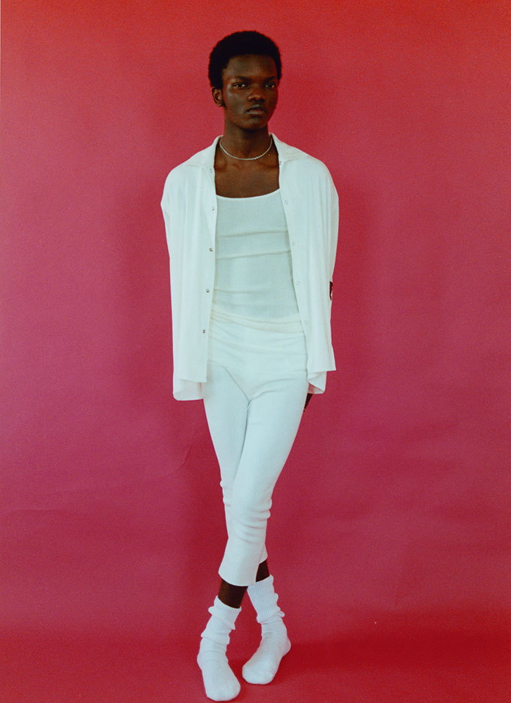Full-body portrait of a Black male model in white clothing against a hot pink background in a studio setting.