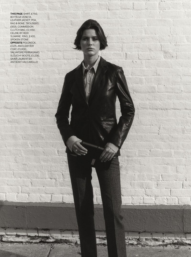 Woman in leather jacket and trousers stands against a brick wall, modeling fashion apparel and accessories for a studio shot.