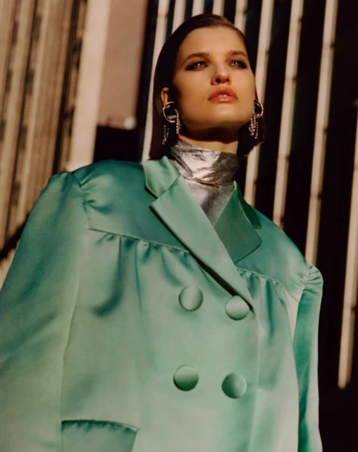 A woman poses in a mint green jacket and silver turtleneck with earrings in an urban setting with vertical bars in the background.