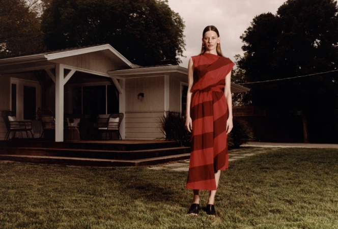 Adult woman in a striped red dress poses outdoors in front of a single-story house with a green lawn in a fashion editorial shoot.