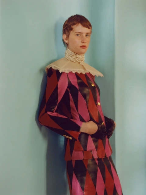 Portrait of a fair-skinned young adult with short auburn hair wearing a harlequin-patterned suit with a pearl necklace against an aqua wall.