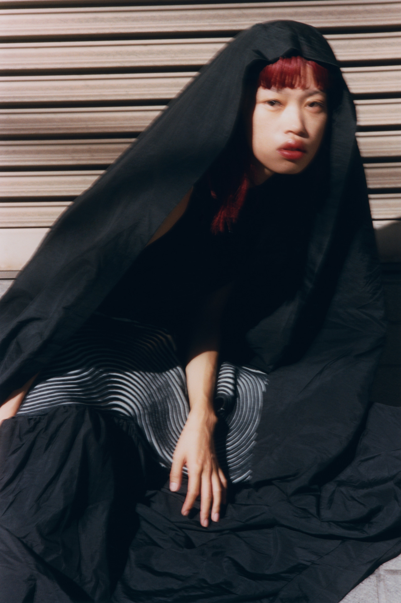 A woman with red hair poses in a black hooded outfit against a horizontally striped surface, creating a striking fashion editorial image.