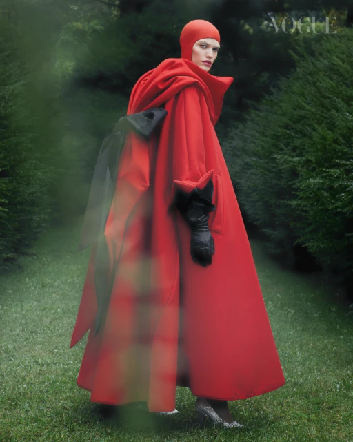 Elegant female fashion model in a red hooded cloak stands in a dreamy green garden. The Vogue logo is visible at the upper right.