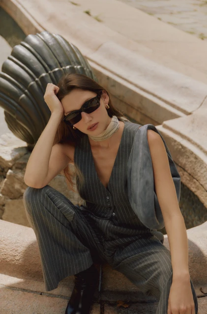 A brunette woman sits in a pinstripe pantsuit with sunglasses in front of a stone structure, bathed in soft light, exuding casual confidence.