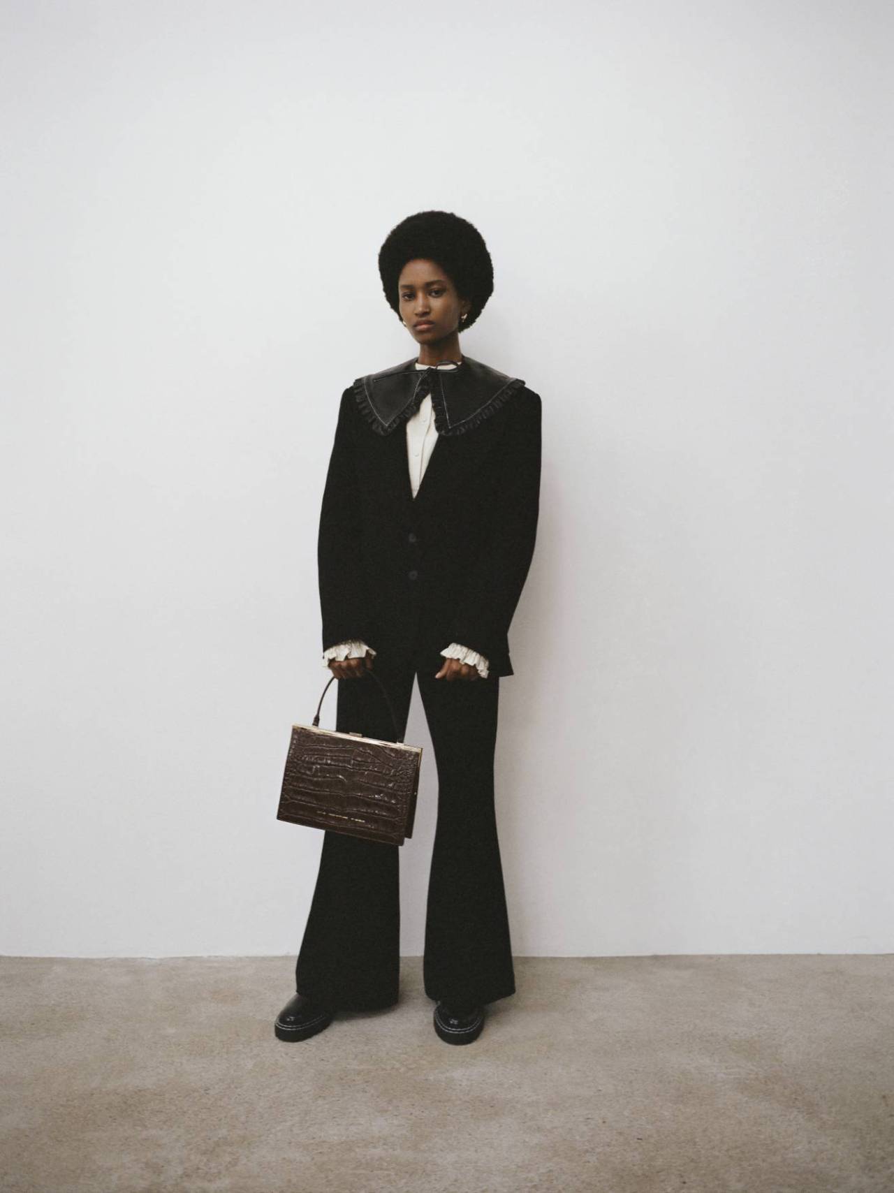 A woman with an afro poses indoors wearing a black suit with wide lapels, ruffled cuffs, and carrying a brown textured handbag.