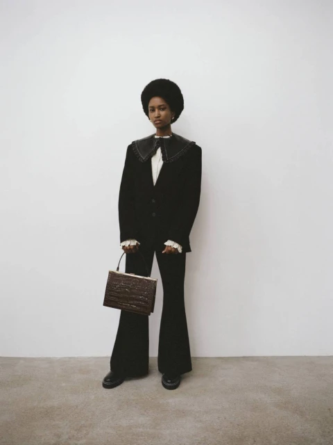 A woman with an afro poses indoors wearing a black suit with wide lapels, ruffled cuffs, and carrying a brown textured handbag.