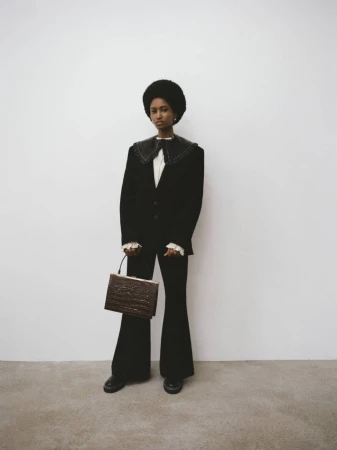 A woman with an afro poses indoors wearing a black suit with wide lapels, ruffled cuffs, and carrying a brown textured handbag.