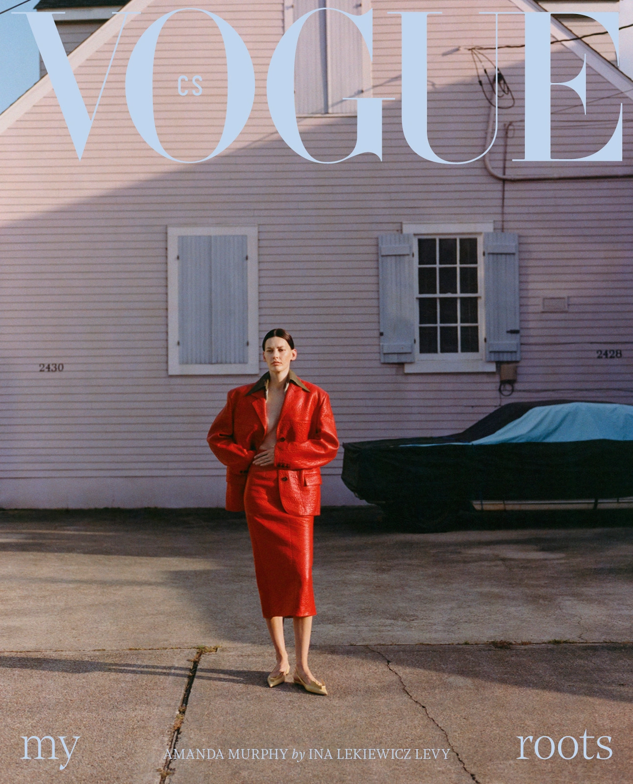 Model wearing a bright red leather suit standing outside a pale pink building with a car parked beside her.