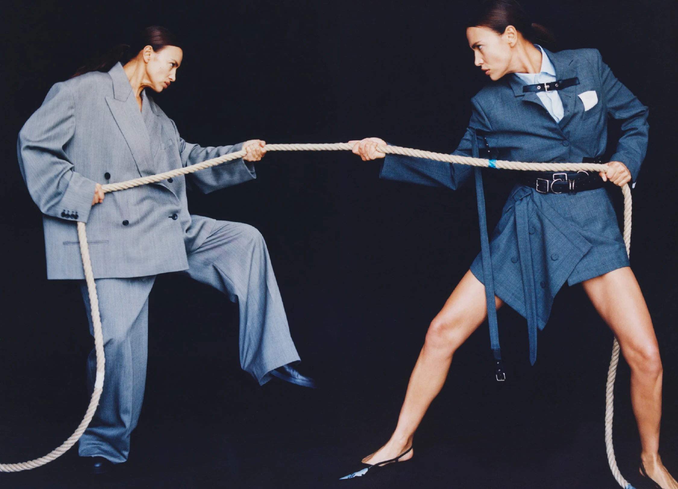 Two women in oversized business suits engage in a tug-of-war with a thick rope, set against a stark black studio backdrop.