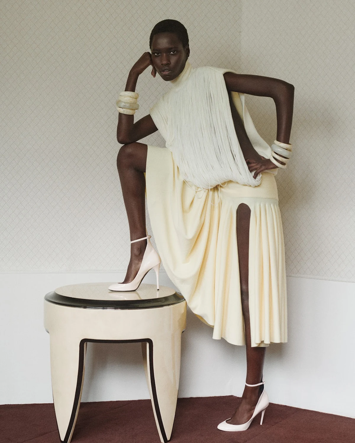 Fashion shot of a single African woman in an elegant cream dress and heels, posing near a table in an indoor setting with soft lighting.