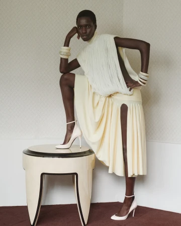 Fashion shot of a single African woman in an elegant cream dress and heels, posing near a table in an indoor setting with soft lighting.