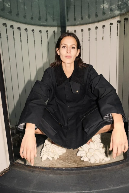 Woman in a black workwear jacket squats indoors wearing sculptural white shoes, looking directly at the viewer with a neutral expression.