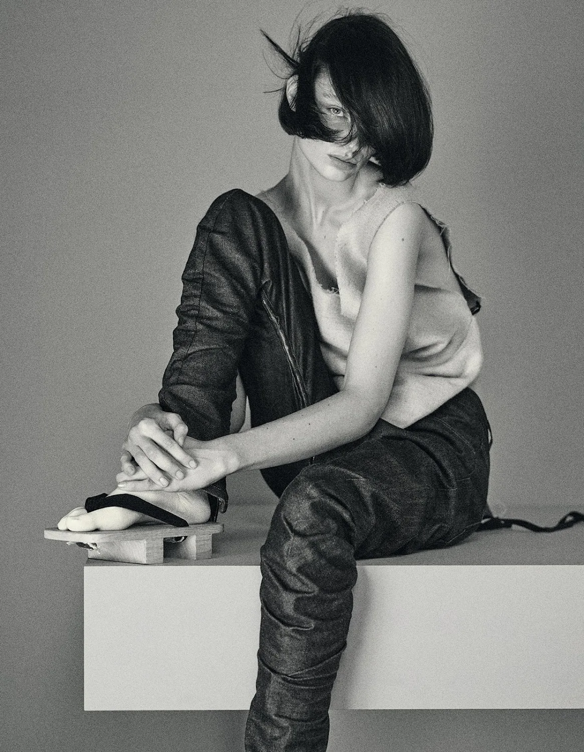A young adult woman with dark hair sits on a platform in a studio, wearing a light-colored sleeveless top, dark-wash denim pants, and platform sandals.