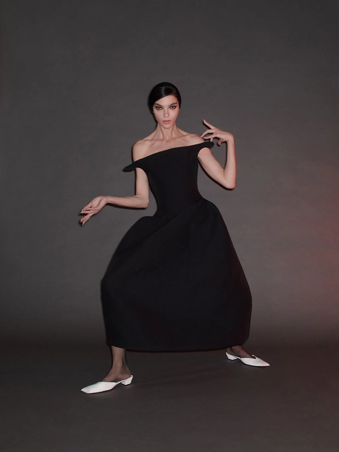Elegant brunette woman in off-shoulder black dress and white flats striking a pose against a grey studio backdrop with soft lighting.