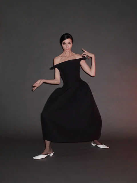 Elegant brunette woman in off-shoulder black dress and white flats striking a pose against a grey studio backdrop with soft lighting.