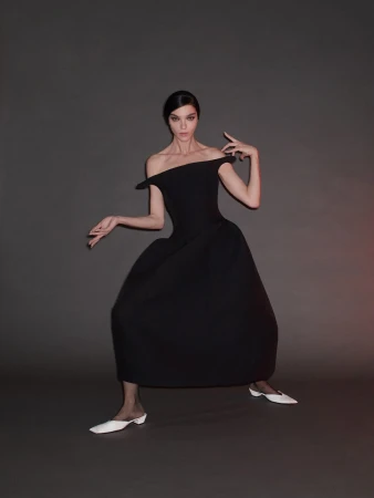Elegant brunette woman in off-shoulder black dress and white flats striking a pose against a grey studio backdrop with soft lighting.