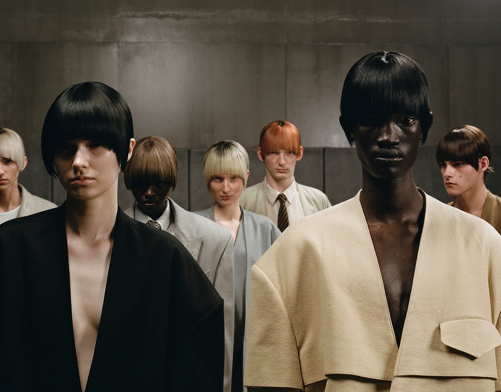 A group of diverse models with uniform bowl-cut hairstyles, posing in neutral-toned clothing against a concrete background, creating a striking visual.