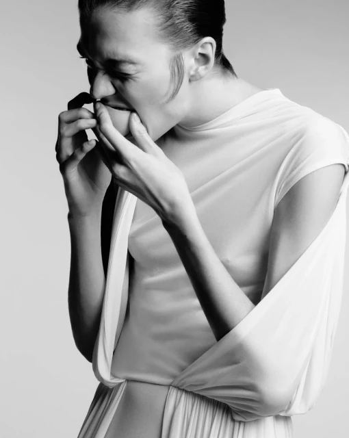 Monochrome portrait of a woman in a white dress with her hand near her mouth, set against a plain, light background.