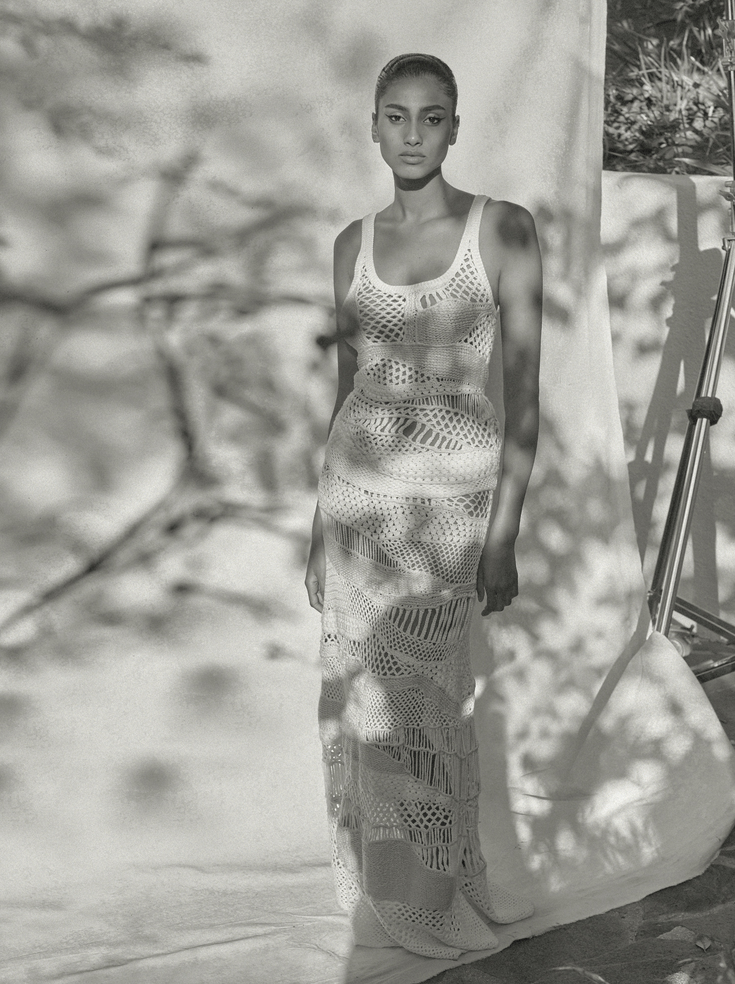 Black and white fashion shot of a woman in a netted dress, posing in front of a backdrop with tree branch shadows.
