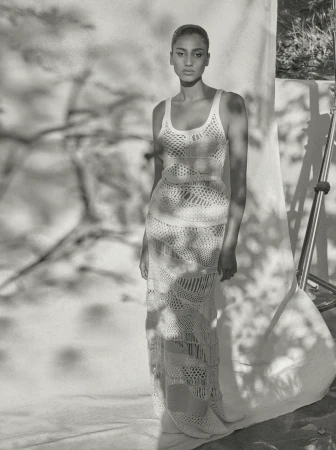 Model in a cream crocheted knit maxi dress standing in front of a white fabric backdrop outdoors.