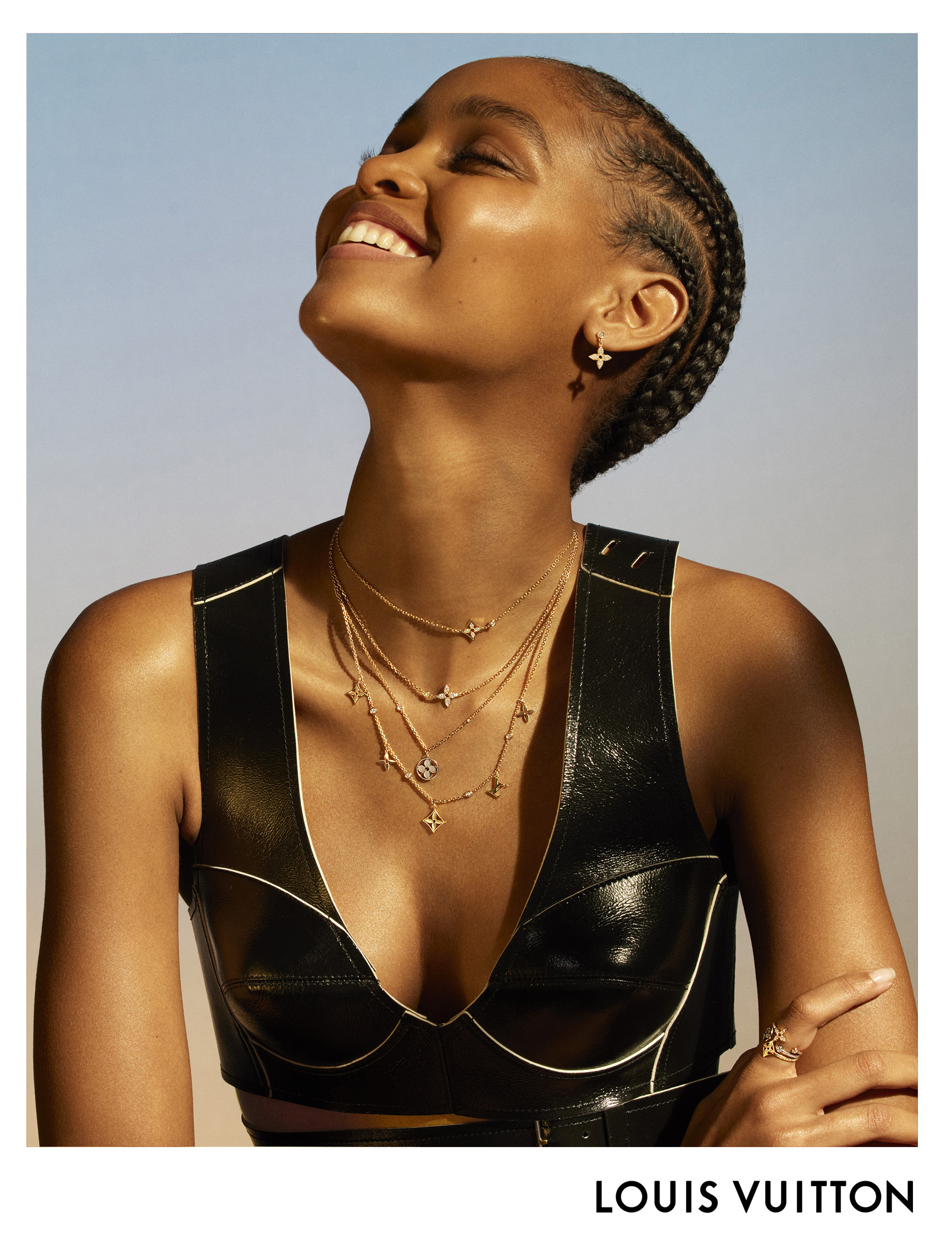 Close-up of a smiling Black woman wearing a black top and layered gold necklaces and earrings, against a light blue background.