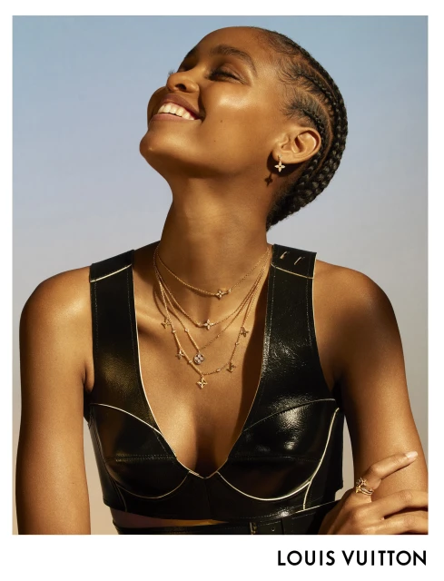 Close-up of a smiling Black woman wearing a black top and layered gold necklaces and earrings, against a light blue background.