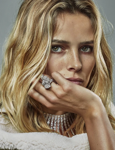 Close-up of a woman with blonde hair wearing a white jacket and a diamond ring and necklace, looking directly at the camera.