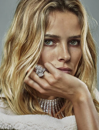 Close-up of a woman with blonde hair wearing a white jacket and a diamond ring and necklace, looking directly at the camera.