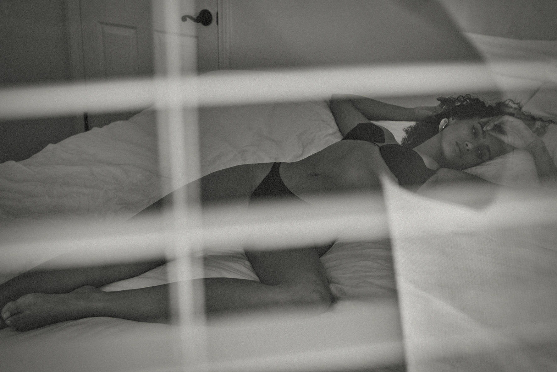 A woman with curly hair lying on a white bed wearing a bra and underwear, partially obscured by window blinds in a grayscale photo.