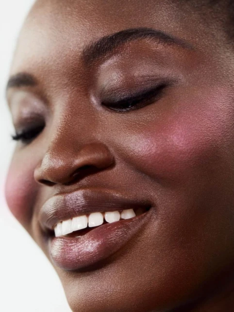 Close-up portrait of a smiling Black woman with elegant eye makeup, pink blush, and glossy lips, set against a clean, bright background.