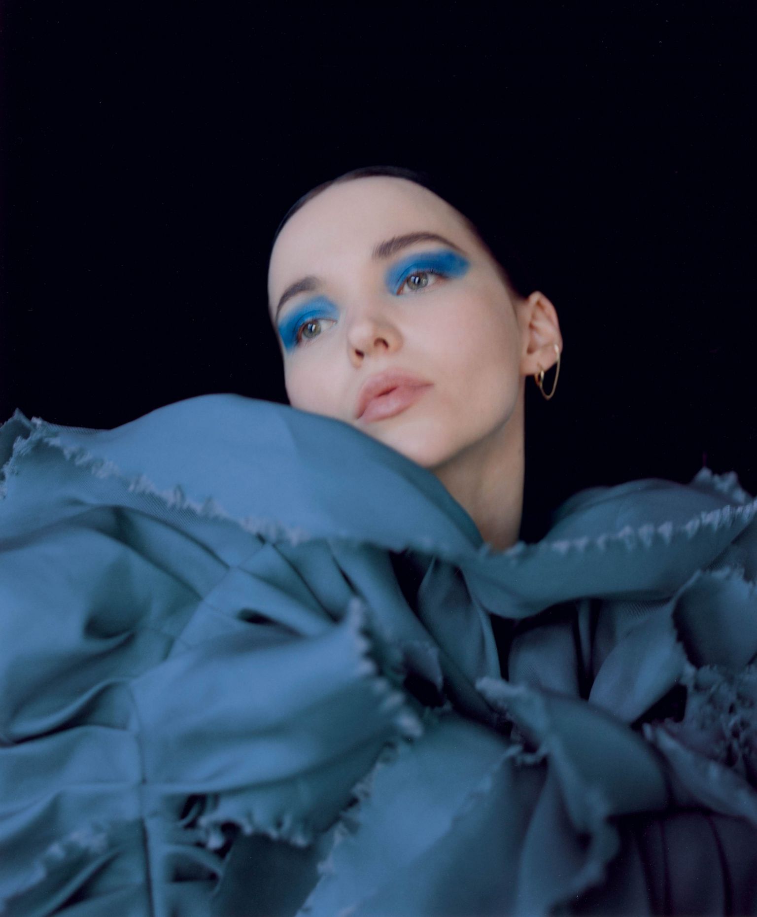 A portrait of a young woman with bright blue eyeshadow, fair skin, and gold hoop earrings posing against a black background.