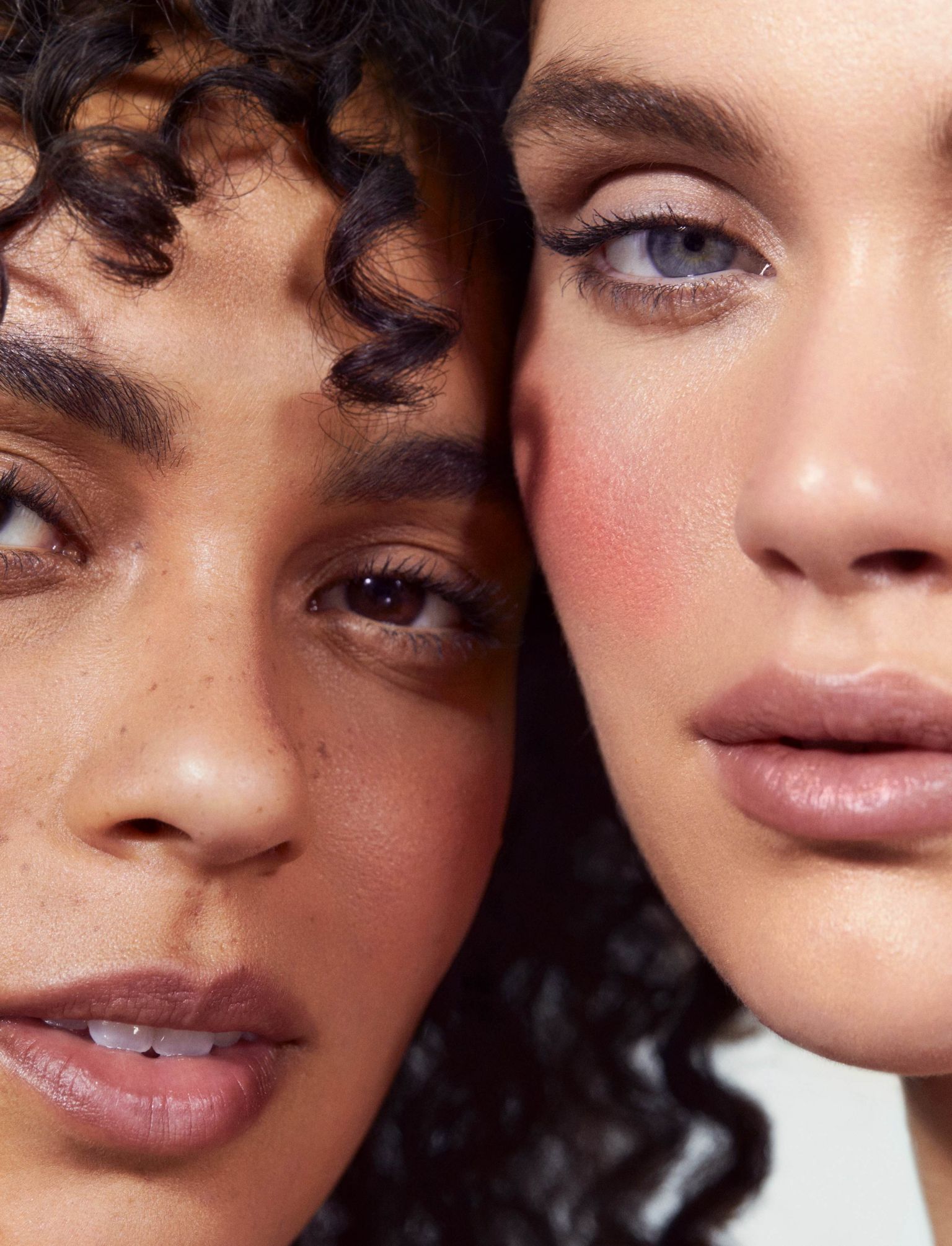 Close-up of two adult women with different skin tones, one with curly hair and the other with straight hair, posing in a beauty shot.