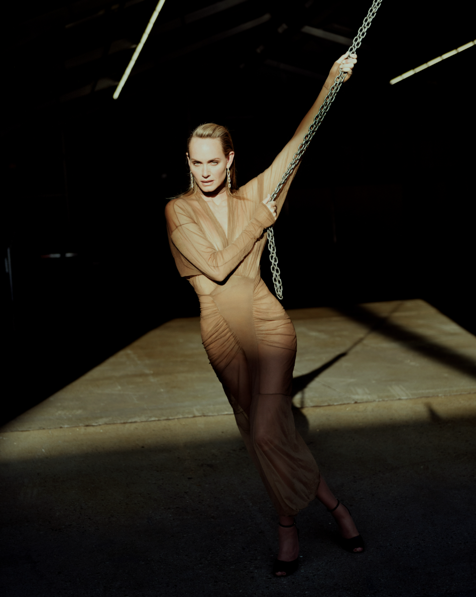 An adult female poses in a brown dress, illuminated by a spotlight in a dimly lit, indoor environment, holding onto a metal chain.