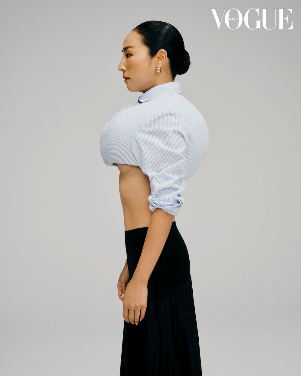 Woman in a profile shot wearing a pinstripe cropped blouse and a black skirt against a solid light gray background in a studio.