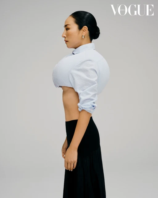 Woman in a profile shot wearing a pinstripe cropped blouse and a black skirt against a solid light gray background in a studio.