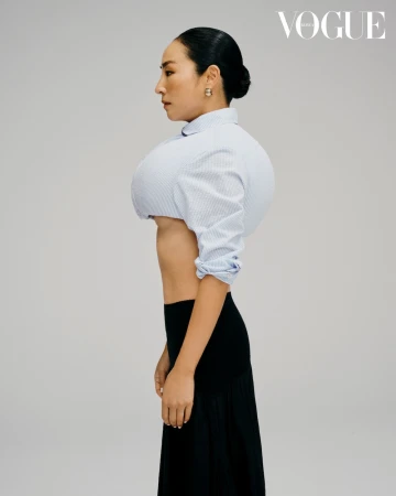 Woman in a profile shot wearing a pinstripe cropped blouse and a black skirt against a solid light gray background in a studio.