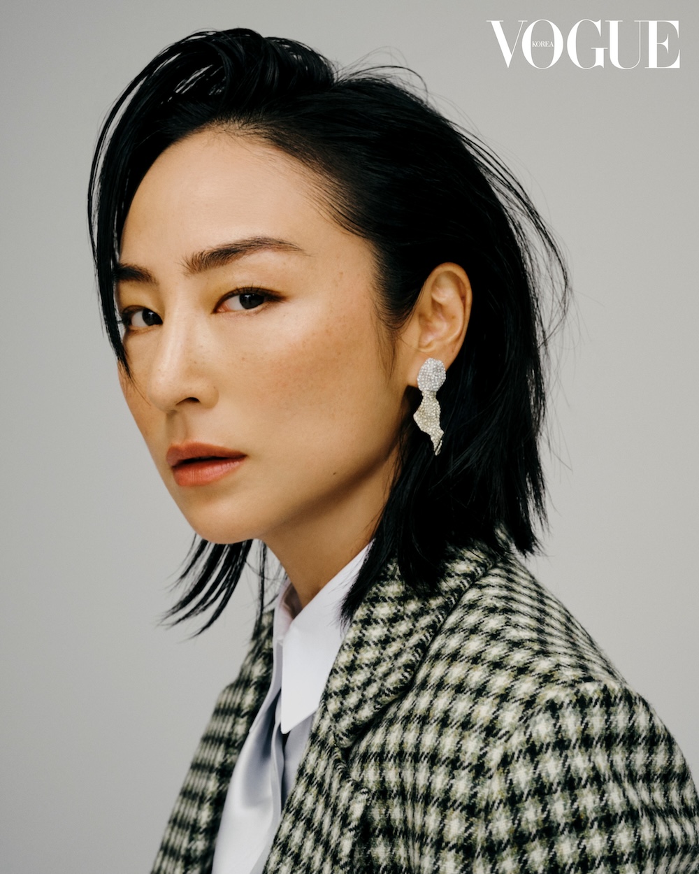 Close-up studio portrait of an Asian woman with black hair wearing houndstooth jacket, white shirt, earrings, neutral studio background, Vogue logo.