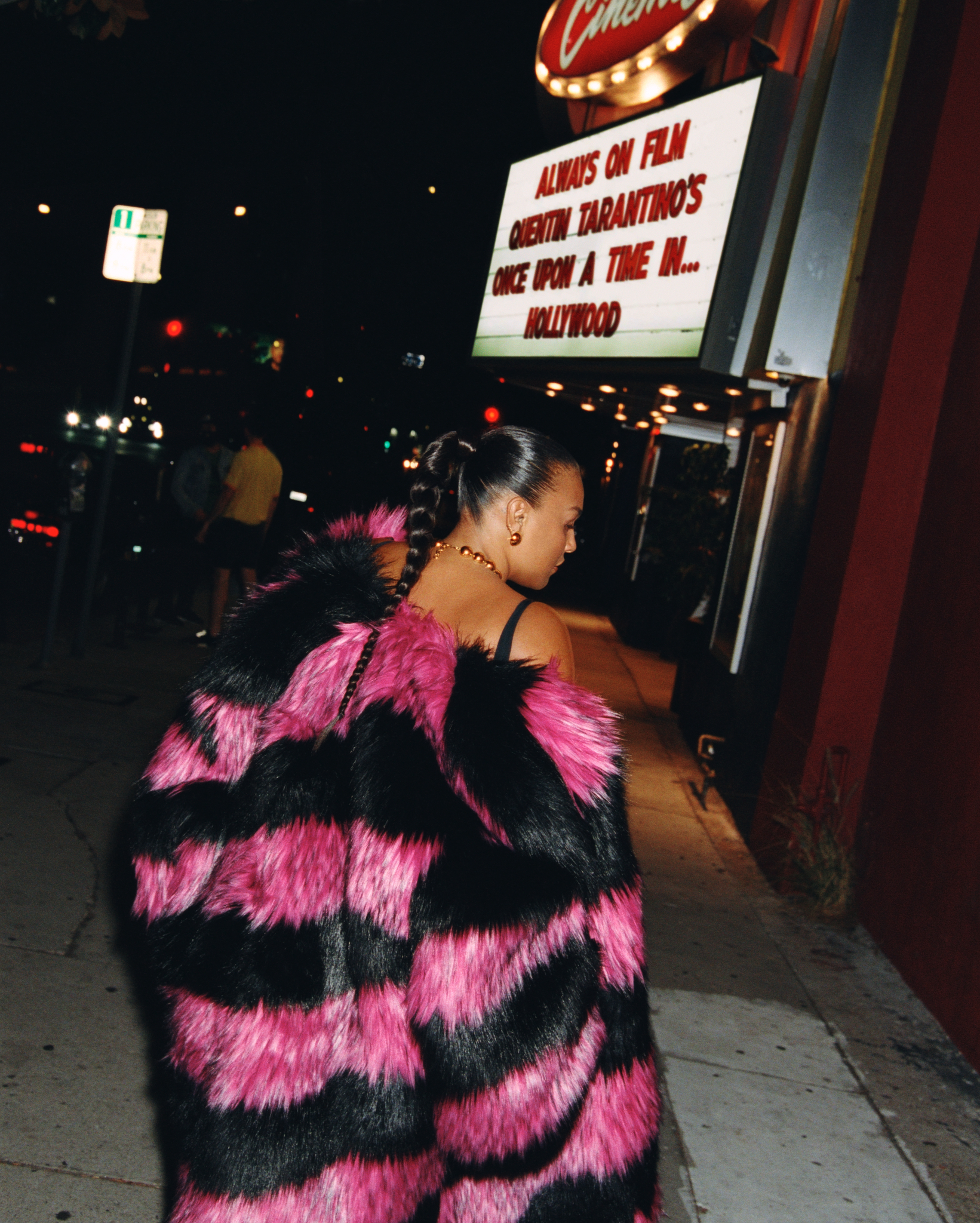 Woman in pink and black striped faux fur coat stands outside a movie theater at night with a lit movie sign in the background.