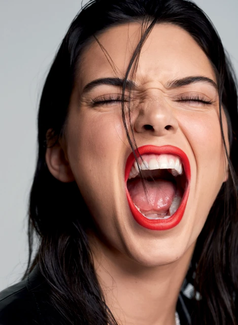 Close-up of an adult female with dark hair, red lipstick, and a black jacket shouting. The shot focuses on her face and expression.