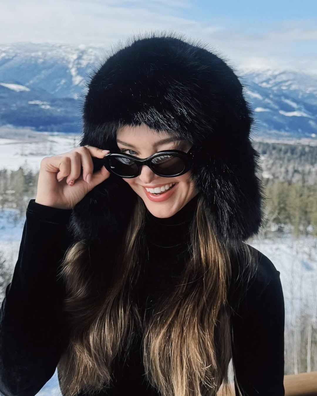 Smiling woman in fur hat and sunglasses poses against a snowy mountain backdrop on a bright day. Fashionable winter portrait.
