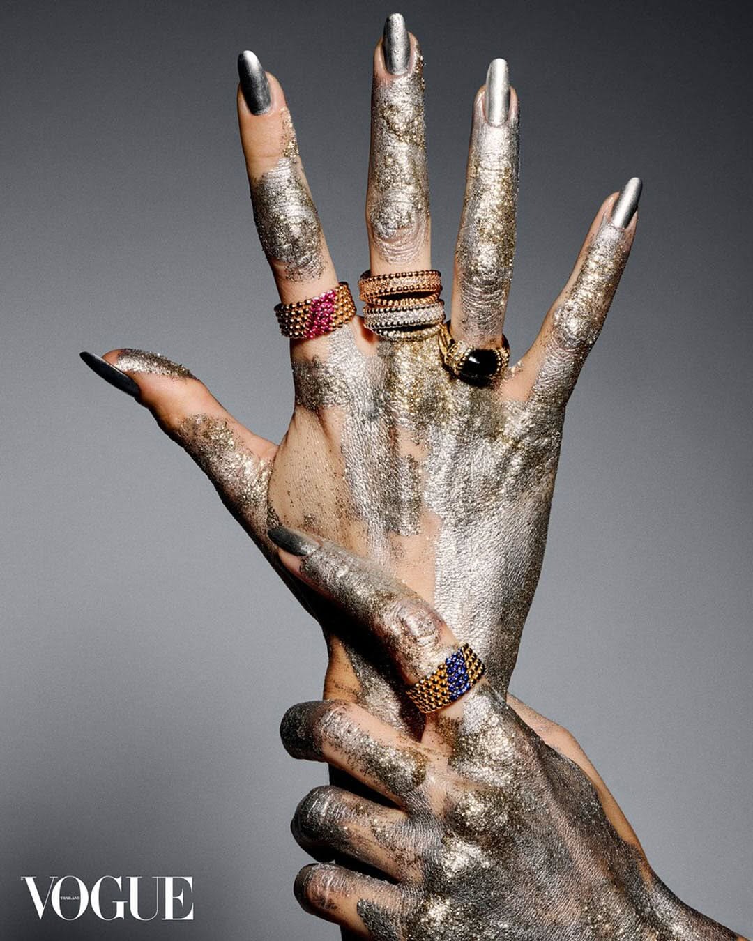 Close-up of hands painted with glitter, showcasing four rings on fingers against a gray background with Vogue magazine logo.
