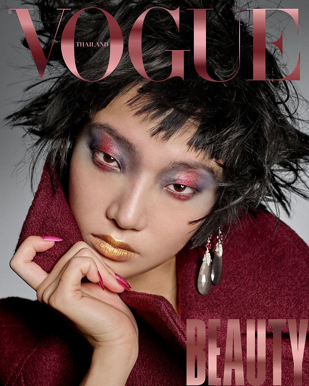 Magazine cover featuring a woman with artistic makeup, statement earrings, and a burgundy coat, overlaid with the 'Vogue Thailand' and 'Beauty' titles.