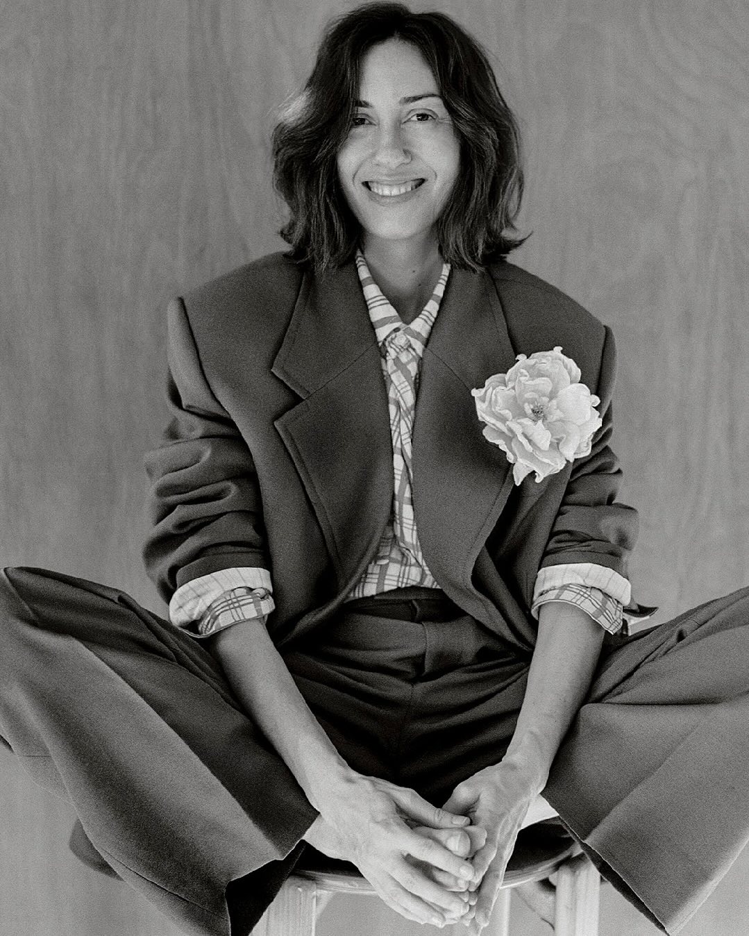 Monochrome portrait of a woman wearing a suit and flower, sitting on a stool. The image is high contrast with a neutral background.