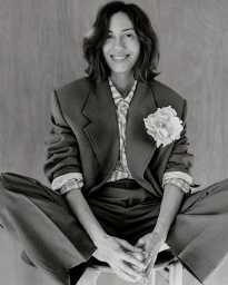 Portrait of a smiling woman wearing an oversized blazer with a lapel flower pinned to it, seated against a wooden wall.