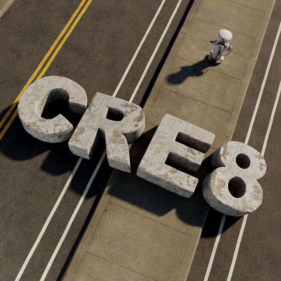 Concrete letters spelling 'CRE8' rest on an asphalt road with a sidewalk, near a white fire hydrant. Overhead shot captures shadows.