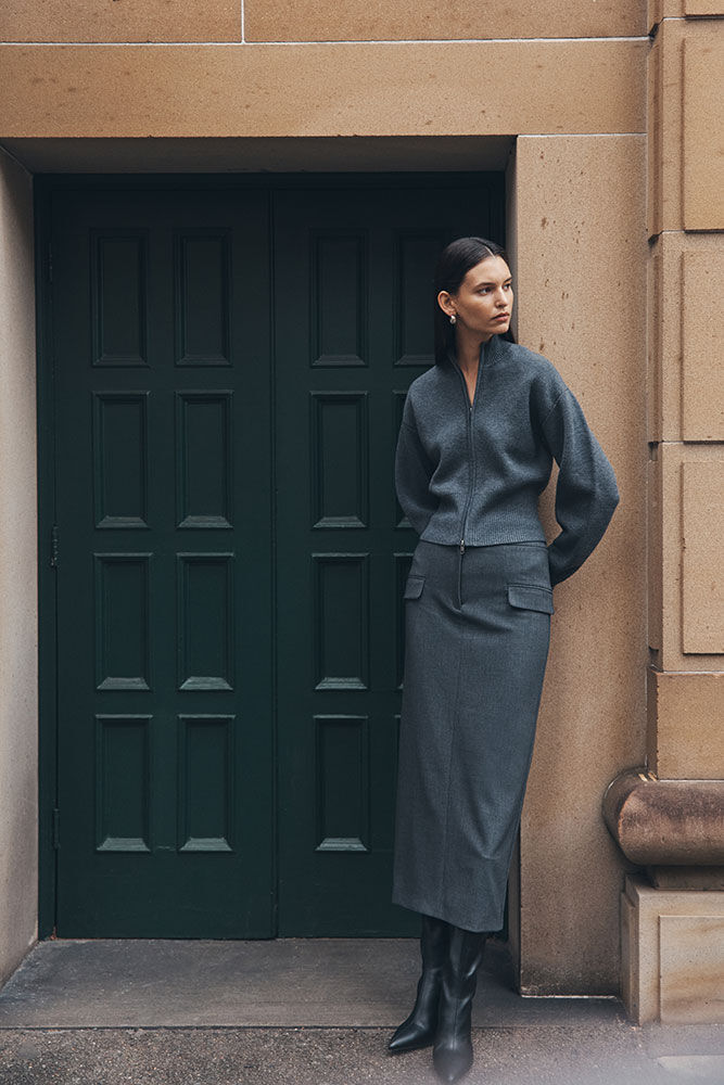 A woman in a grey skirt suit and black boots stands in front of a dark green paneled door set in a tan stone building facade.