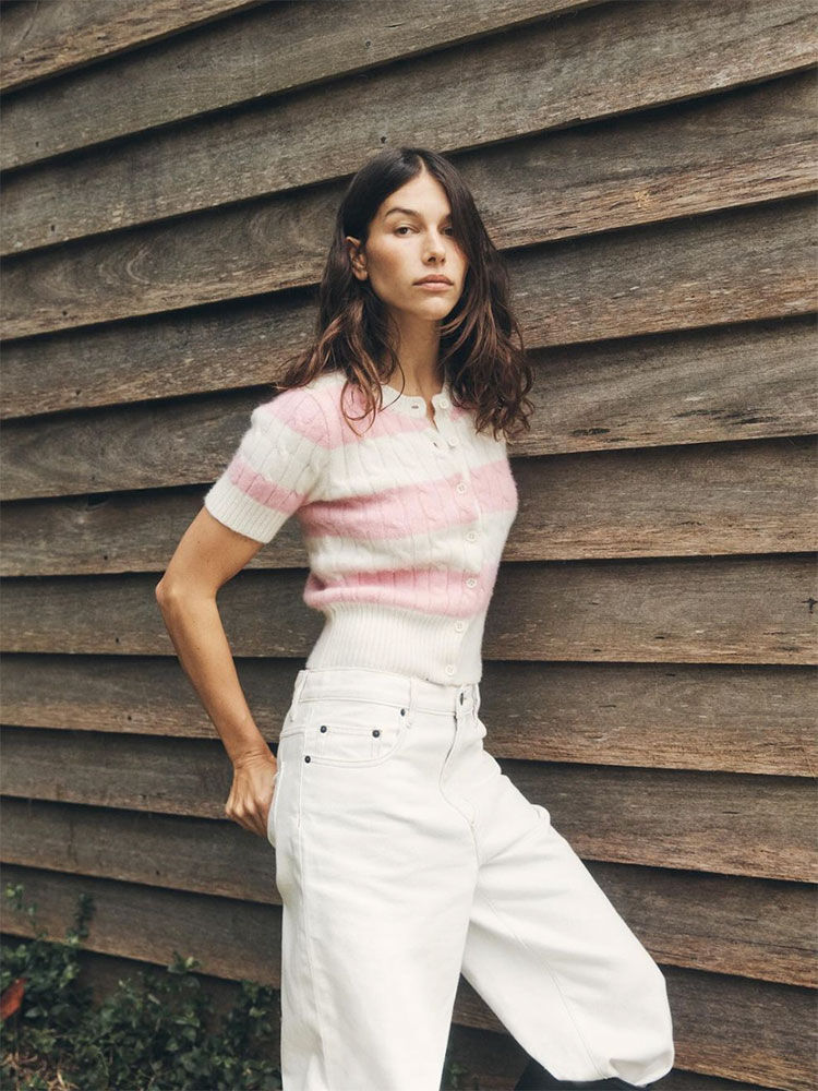 A woman stands against a wooden wall, wearing a striped pink and white sweater and white pants in a medium shot.