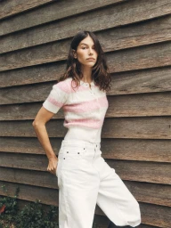 Woman in short sleeve pink and white striped cardigan and white jeans posing against wooden slat wall
