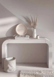 Minimalist white console table with a decorative bowl, stack of books, and vase of dried grass against a neutral wall.