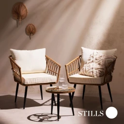 Two woven rattan armchairs with cream cushions and a small round side table arranged against a beige wall with hanging fans